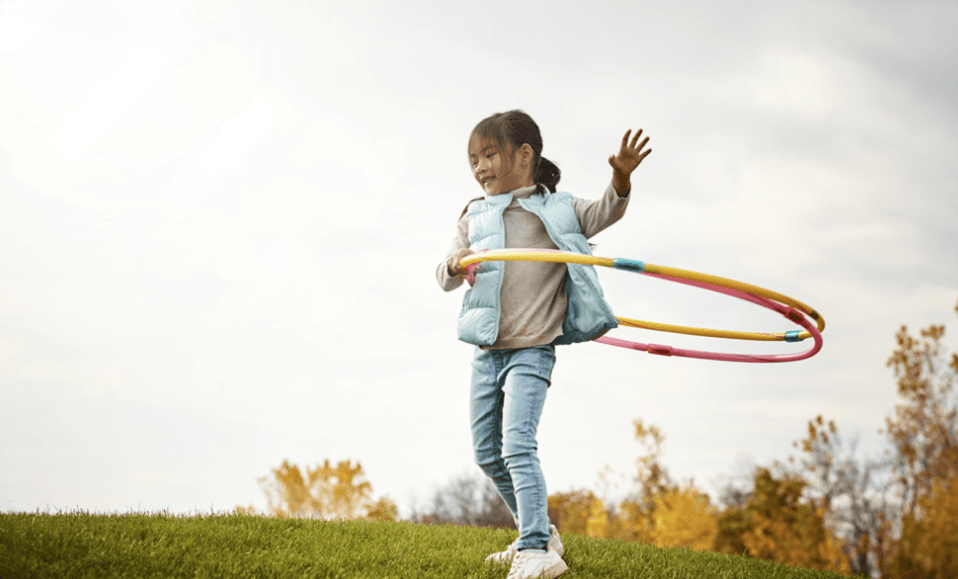 A girl spinning a hula hoop on a grassy hill under fall trees